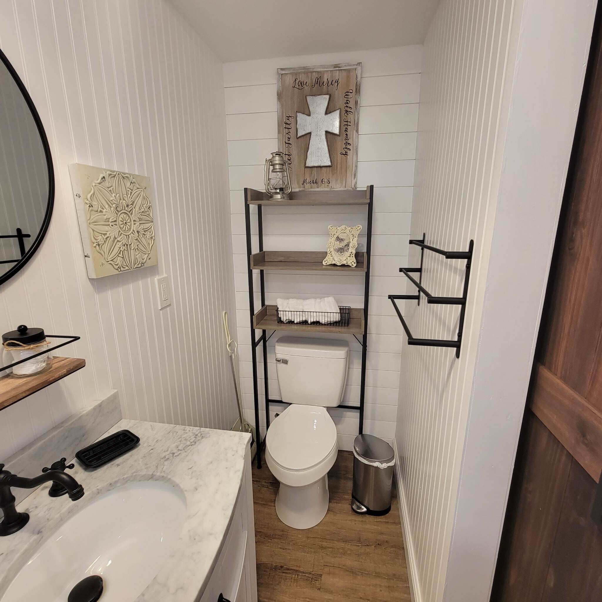 Une petite salle de bain avec un lavabo blanc, un miroir, des toilettes et une étagère décorée d’une croix.