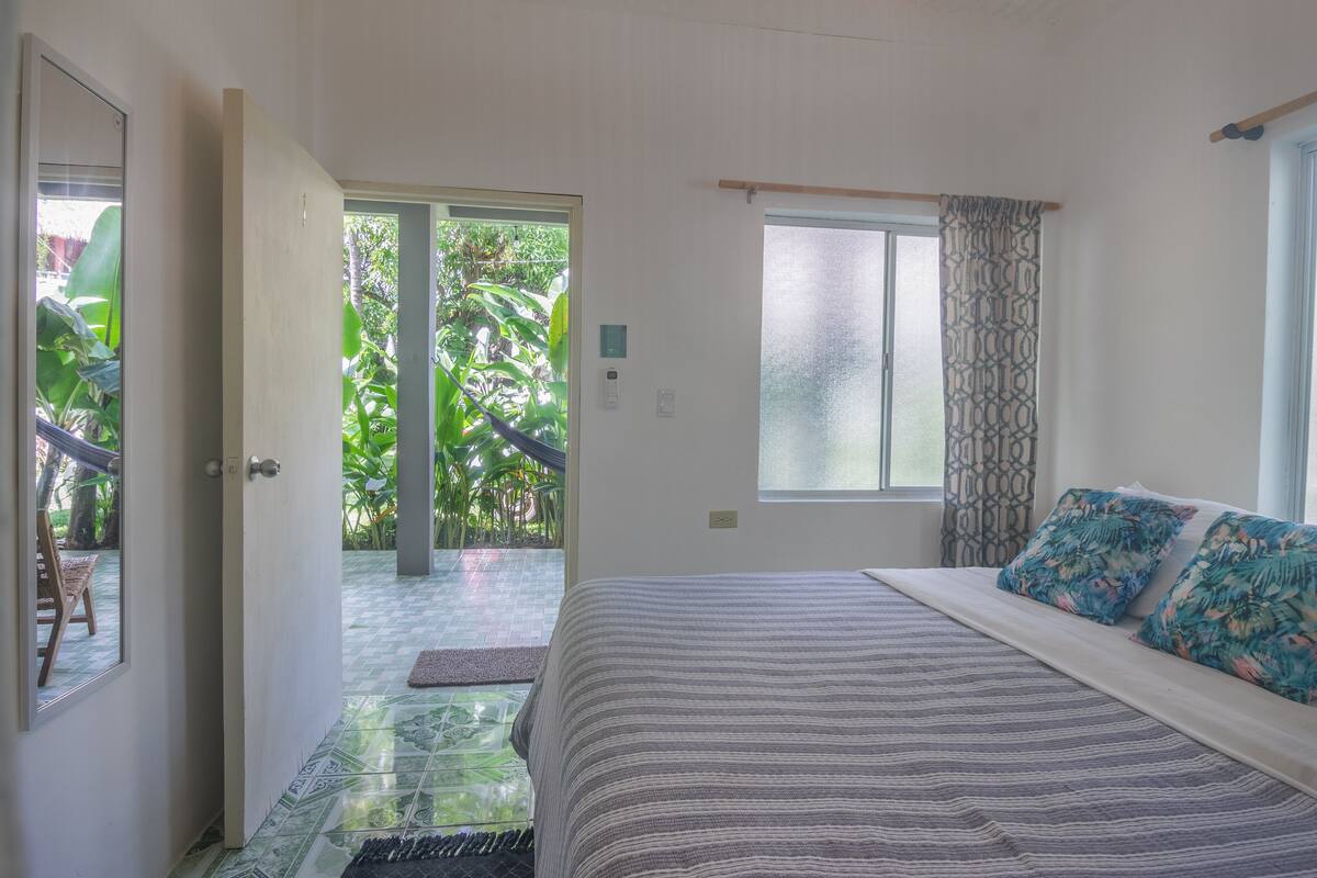 Un dormitorio con cama, una ventana con cortinas estampadas y una puerta que da a un balcón con vistas a la vegetación.