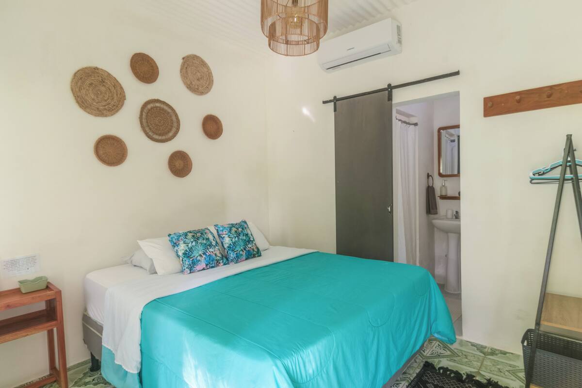 Un dormitorio con una cama, una mesita de noche de madera, un aire acondicionado de pared y una puerta que da al baño.