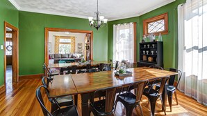 Een eetgedeelte met een houten tafel en stoelen, een kroonluchter, en uitzicht op een woonkamer met een bank en gordijnen.