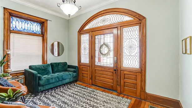 Een woonkamer met een groene bank, een rug met patroon, een houten deur met glas-in-lood en een kroonluchter.