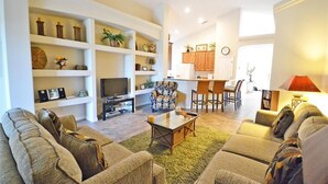 Una sala de estar con un sofá, una mesa de centro y un televisor. Hay una cocina con armarios de madera y una mesa de comedor con sillas.