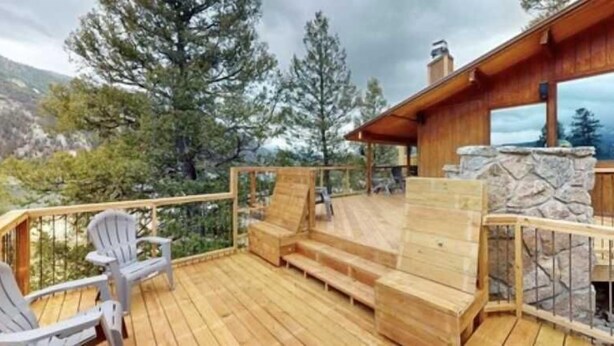 Une terrasse en bois avec des fauteuils Adirondack, une cheminée en pierre et une vue sur les arbres et les montagnes.