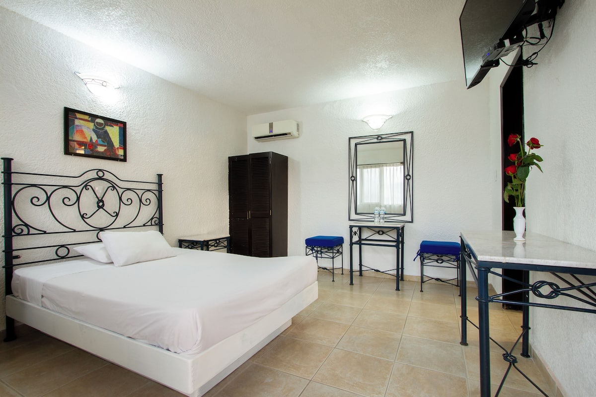 Un dormitorio con cama, armario, escritorio, silla, espejo y un florero con flores.
