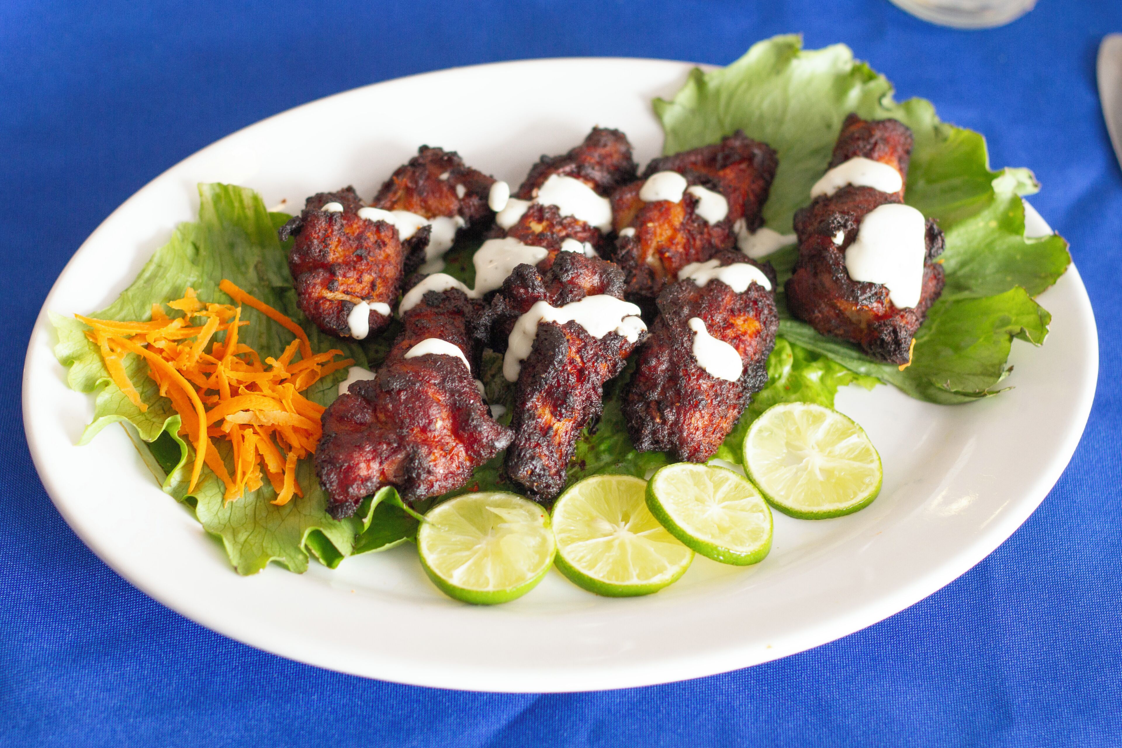 A plate of grilled meat skewers with a white sauce, garnished with shredded carrots and lime slices, on a white plate.