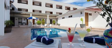 Zona de piscina con sillones de descanso, una mesa con bebidas y un edificio con varios balcones.