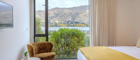 A hotel room with a bed, a chair, a small table, and a view of a lake and mountains.