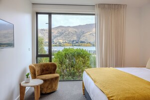 Habitación de hotel con cama, una silla, una mesita y vistas a un lago y montañas.