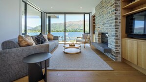 A modern living room with a sofa, coffee table, and stone fireplace. Large windows offer a view of a lake and mountains. - Marina Terrace Apartments (Wanaka)