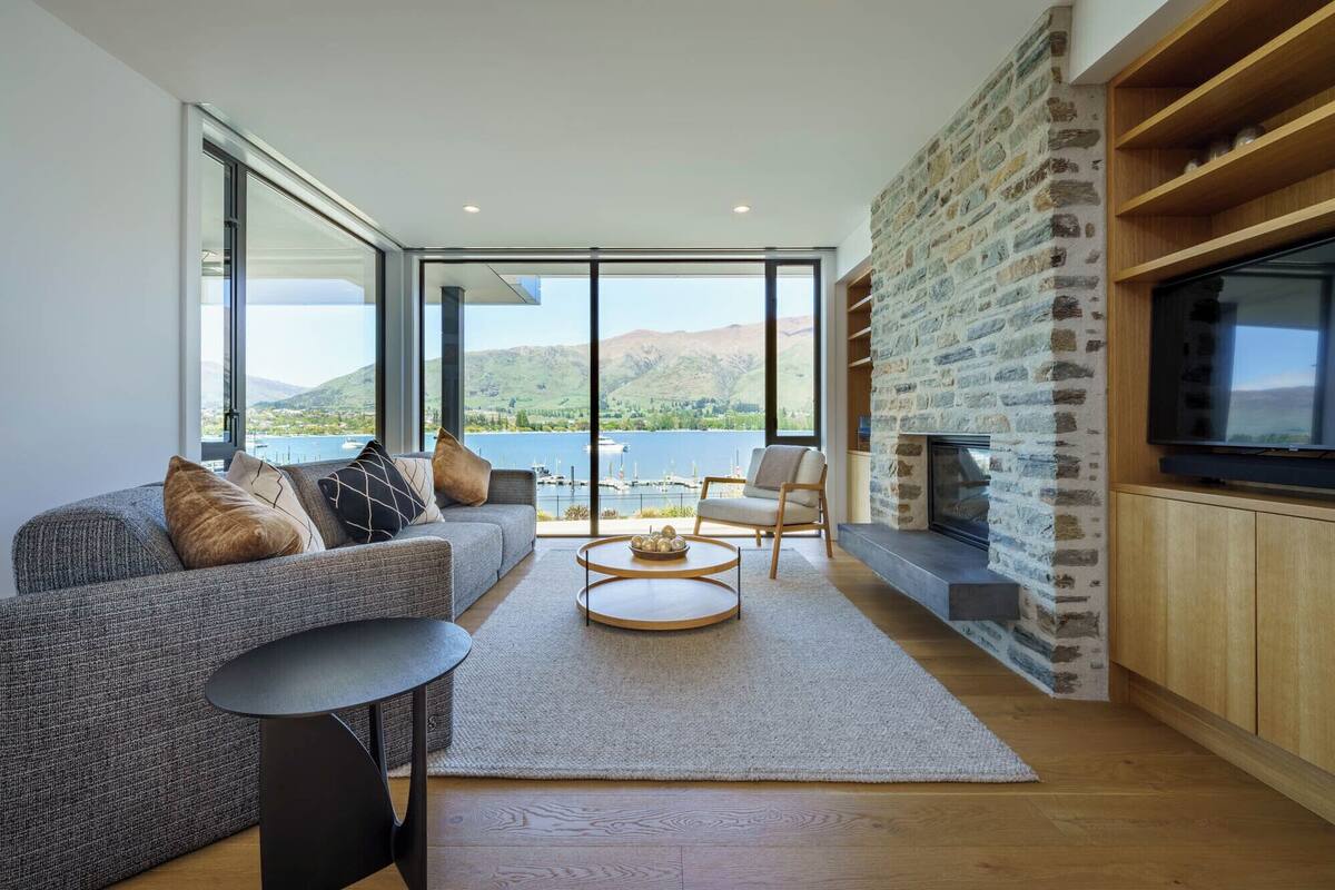A modern living room with a sofa, coffee table, and stone fireplace. Large windows offer a view of a lake and mountains.