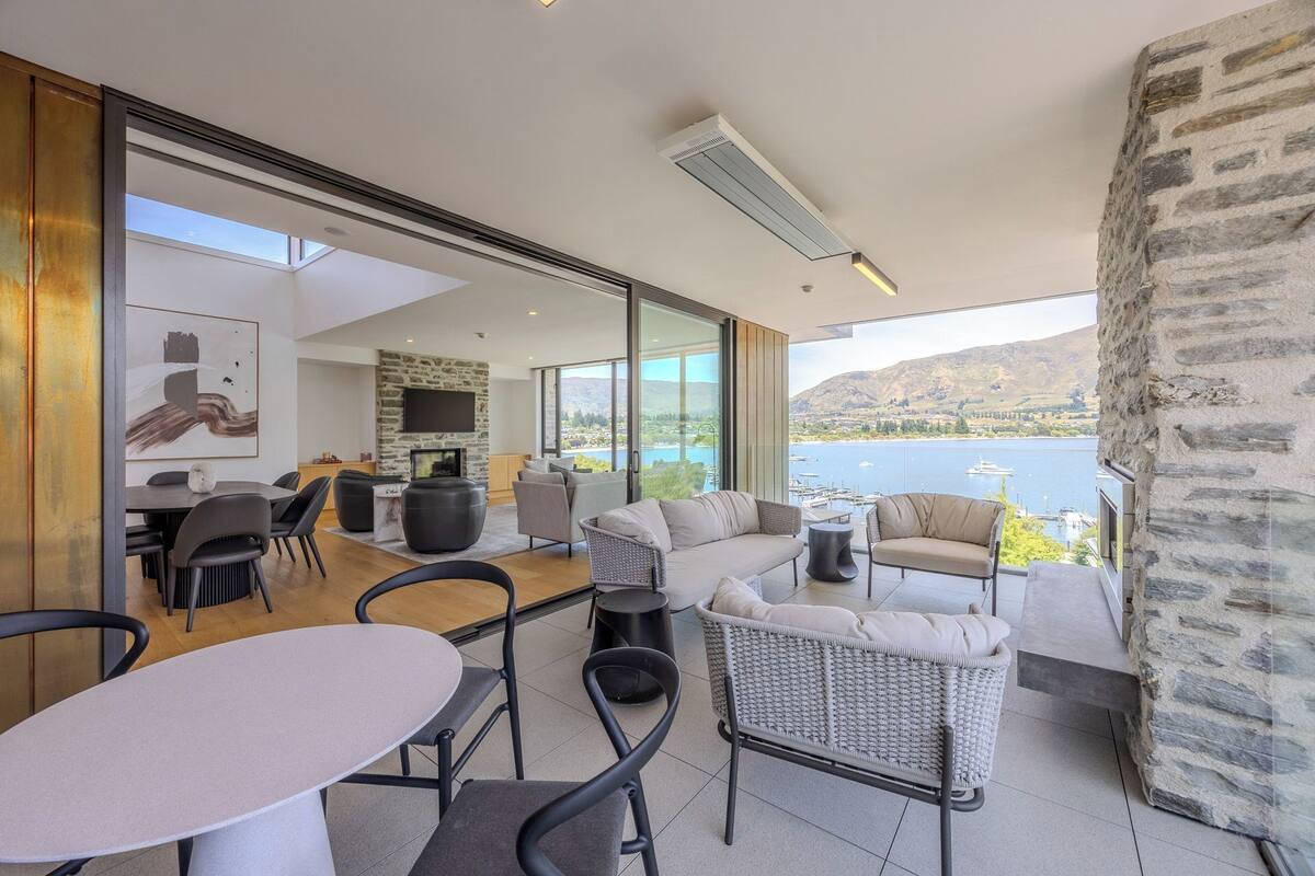 A modern living room with a stone fireplace, a dining area, and a balcony with a view of a lake and mountains.