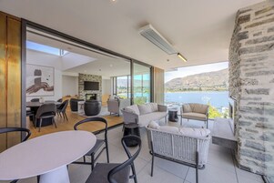 A modern living room with a stone fireplace, a dining area, and a balcony with a view of a lake and mountains.