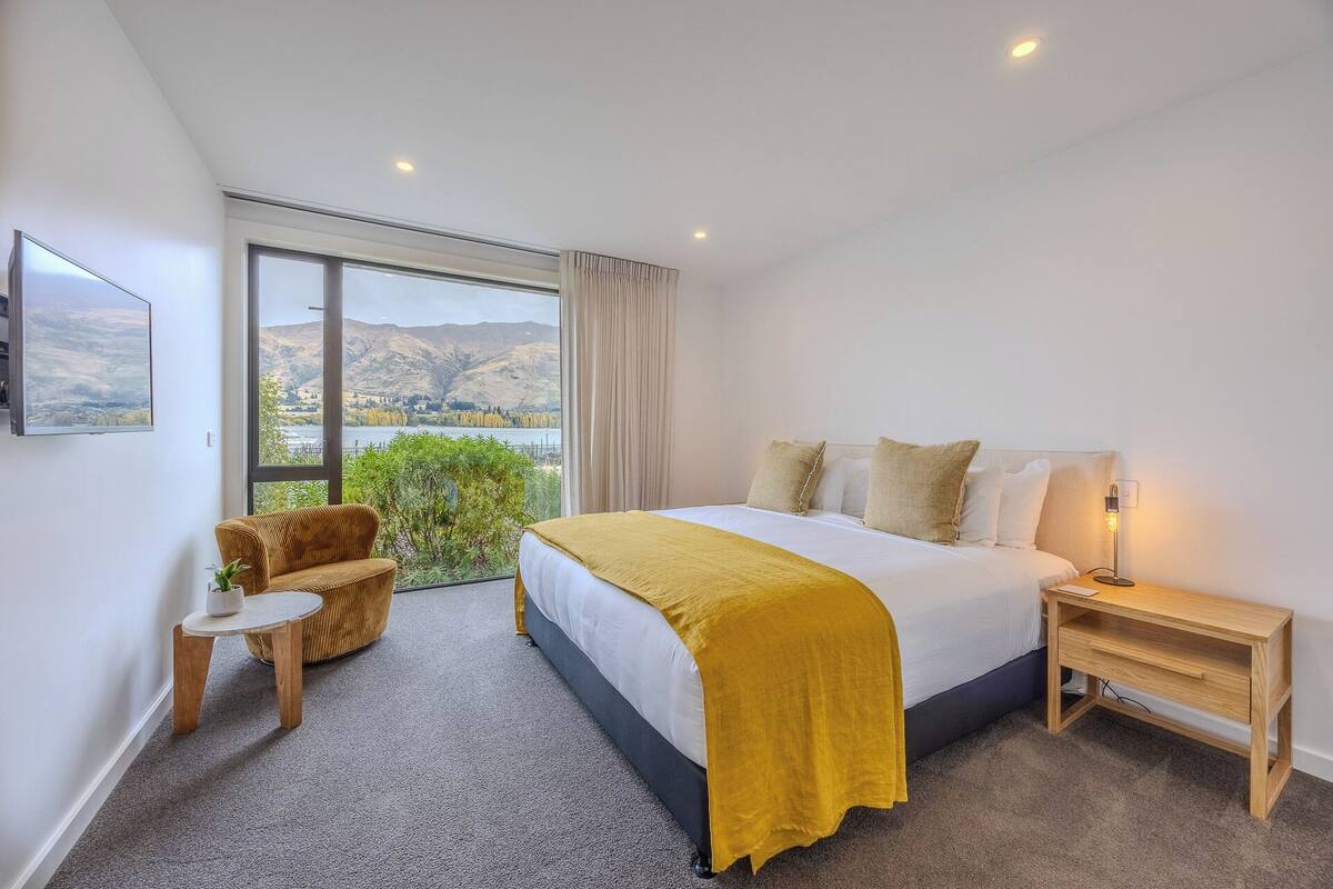 A bedroom with a large window, a bed with a yellow throw, a bedside table with a lamp, and a chair.