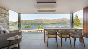 A modern room with a large window overlooking a lake and mountains, furnished with a sofa, table, and chairs. - Marina Terrace Apartments (Wanaka)