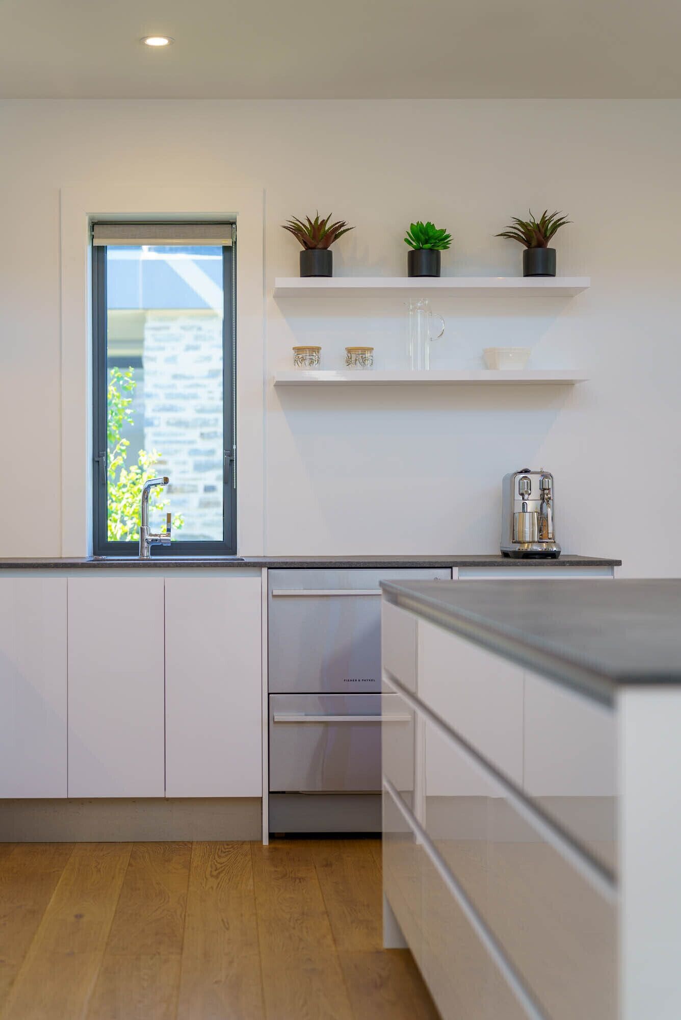 Una cocina moderna con armarios blancos, lavavajillas integrado, cafetera y ventana con vistas al exterior.