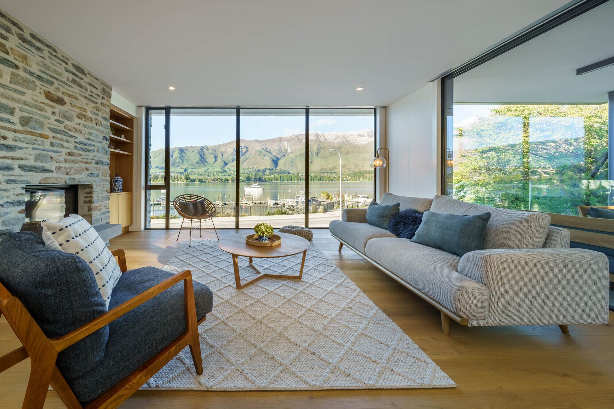 Ein modernes Wohnzimmer mit einer Steinwand, einer Couch, einem Sessel, einem Couchtisch und Blick auf einen See und Berge durch große Fenster.