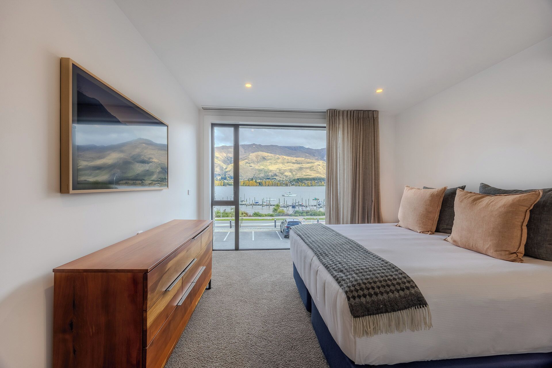 Ein modernes Schlafzimmer mit einem großen Bett, einer Holzkommode und einem großen Fenster mit Blick auf einen See und Berge.