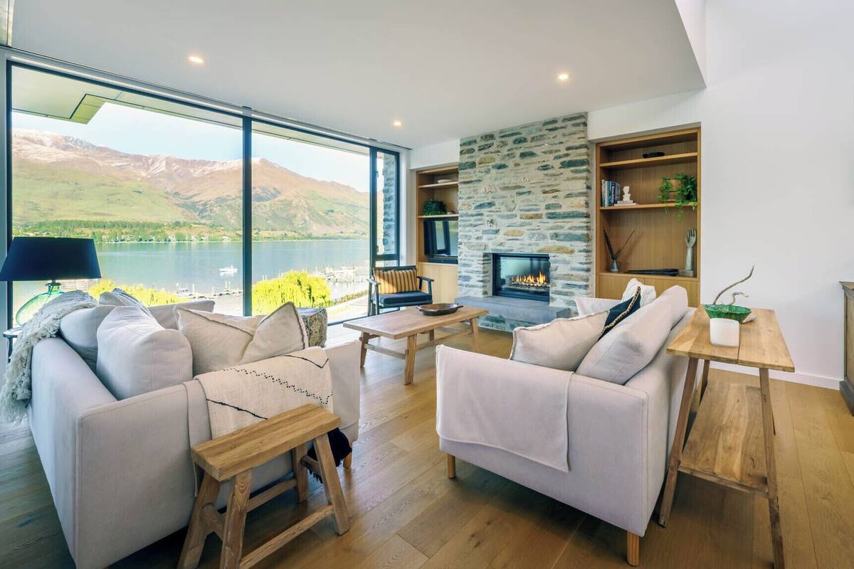 A modern living room with a stone fireplace, large windows overlooking a lake, and a cozy seating area.