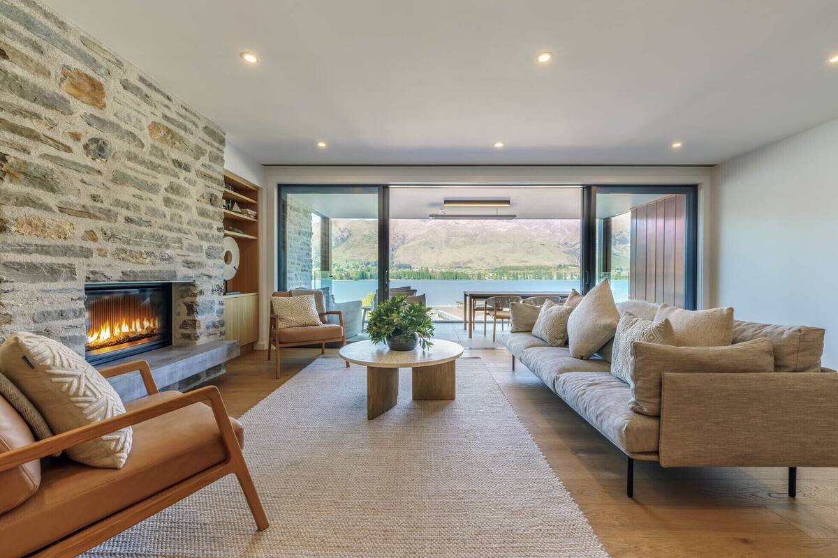 A modern living room with a stone fireplace, comfortable seating, and a view of a lake and mountains.