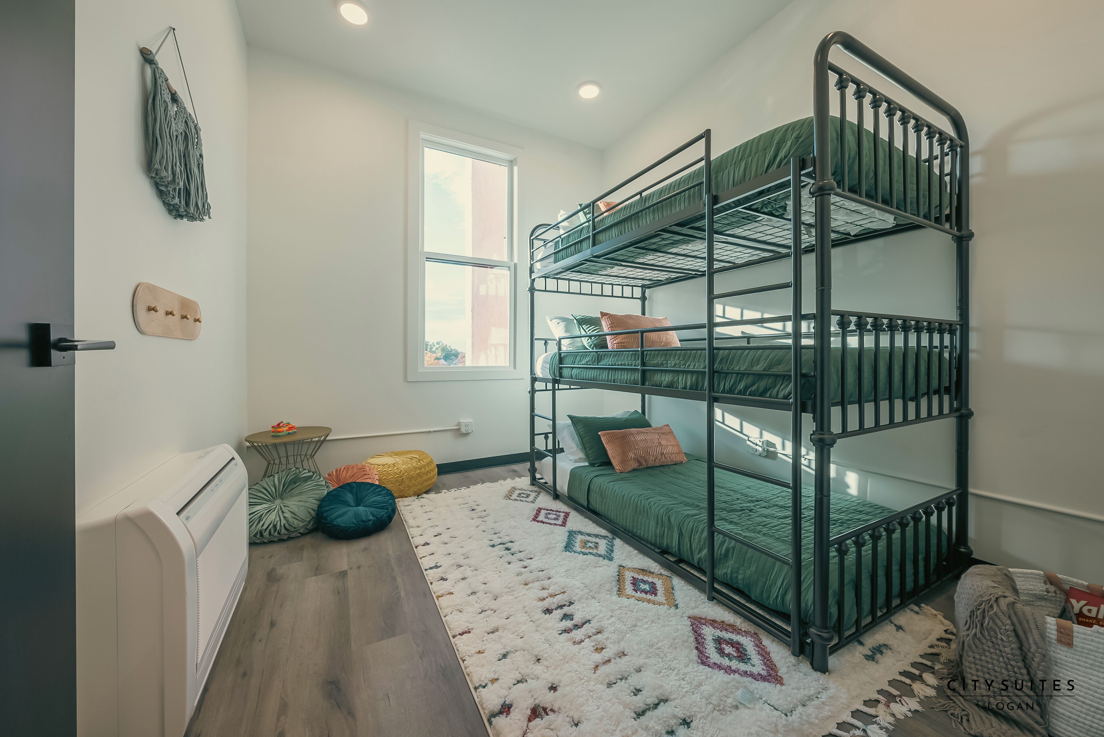 Une chambre avec un lit superposé, une fenêtre, un radiateur, un tapis à motifs et une petite table avec des coussins.