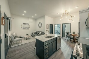 Cozinha moderna com ilha central, sala de jantar com mesa e cadeiras de madeira e sala de estar com sofá e mesa de centro.