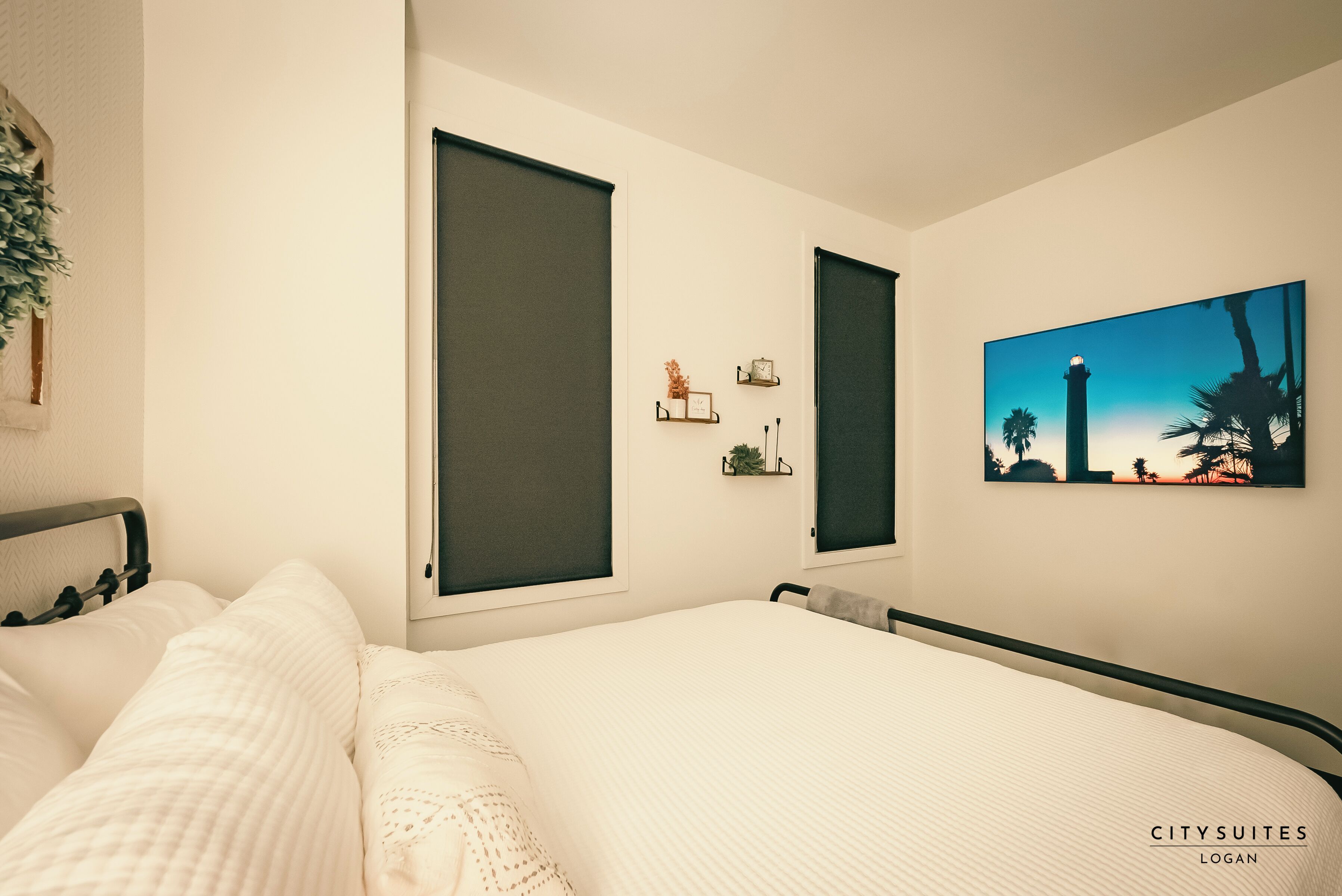 A bedroom with a large bed, two windows, a wall-mounted clock, and a framed picture of a lighthouse.