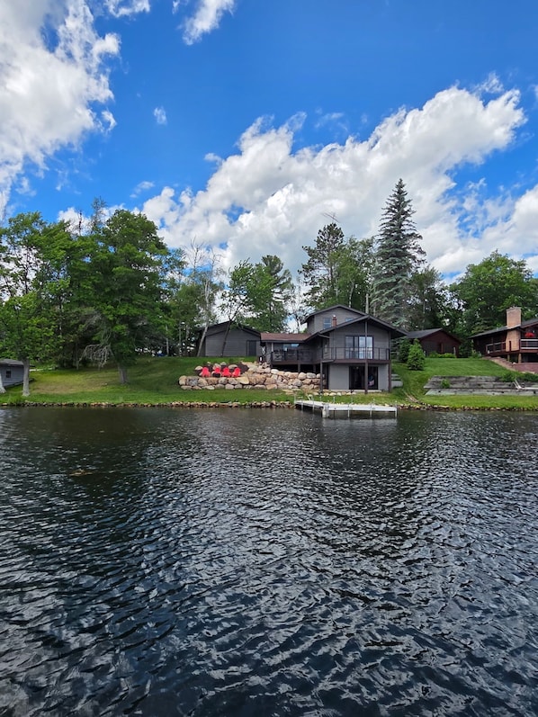 A lakeside property with a dock, a stone wall with red chairs, and a large house with a garage.