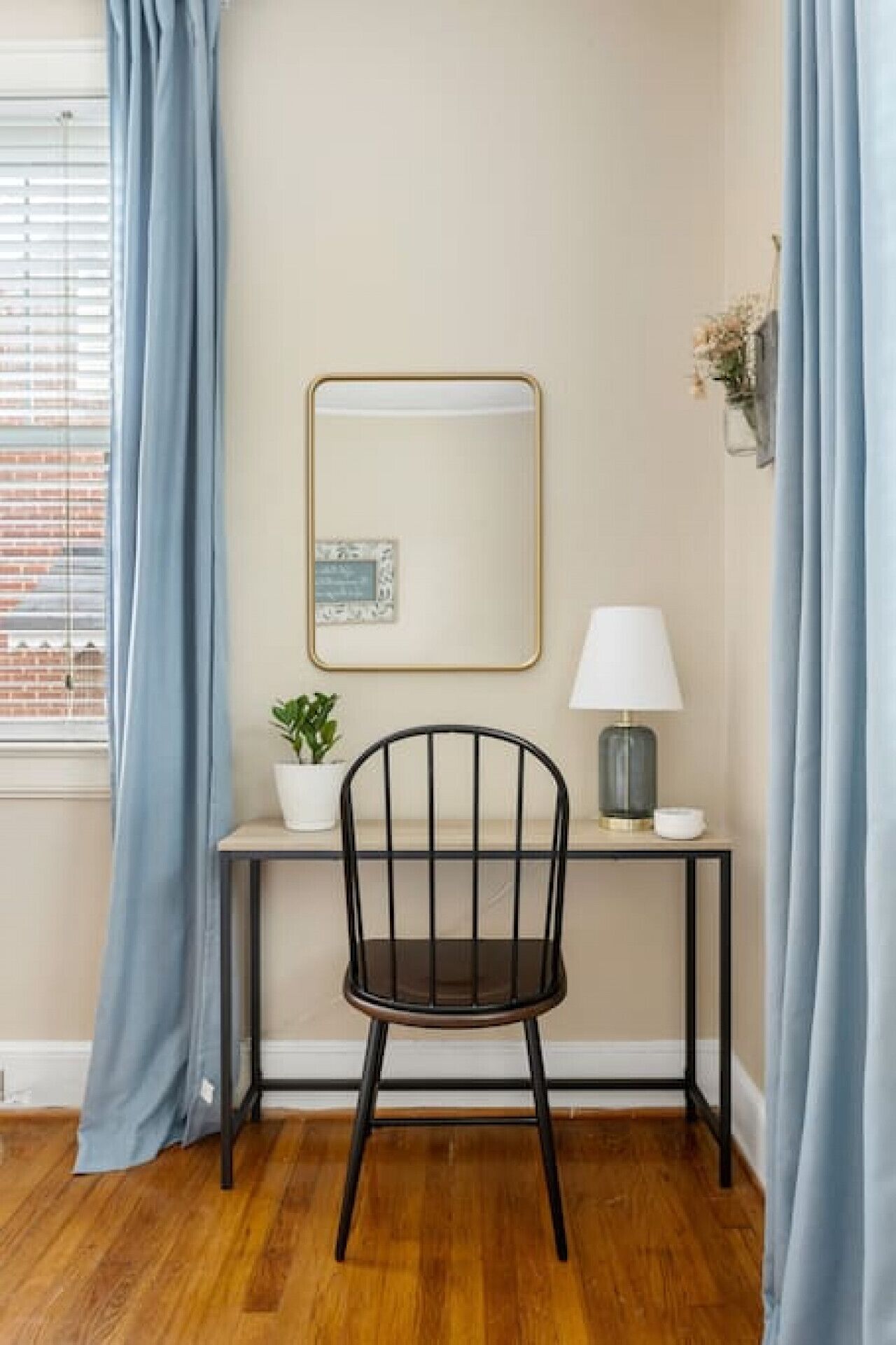 Ein Zimmer mit Holzboden, Schreibtisch mit Stuhl, Lampe, Zimmerpflanze und einem gerahmten Bild an der Wand.
