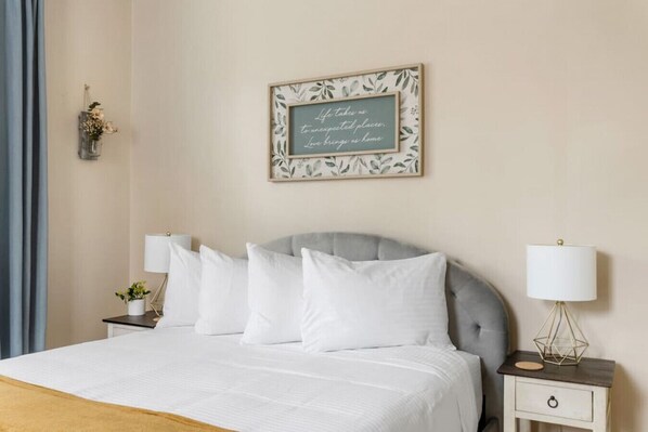 A neatly made bed with white linens, a grey upholstered headboard, and a bedside table with a lamp. A framed quote hangs on the wall above the bed.