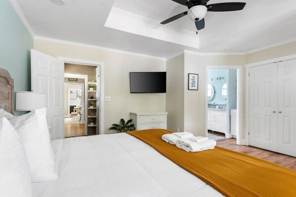 Habitación de hotel con cama, cabecera de madera, ventilador de techo, televisor montado en la pared y baño visible a través de una puerta abierta.