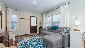 A living room with a grey sectional sofa, a patterned rug, a white side table, and a door with a cross on the wall.