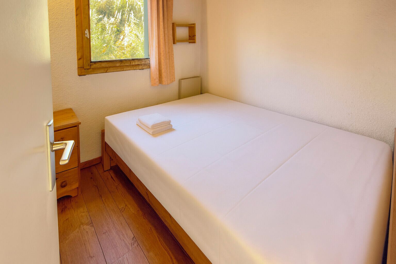 A small, tidy room with a single bed, a wooden nightstand, a window with curtains, and a wall-mounted light.