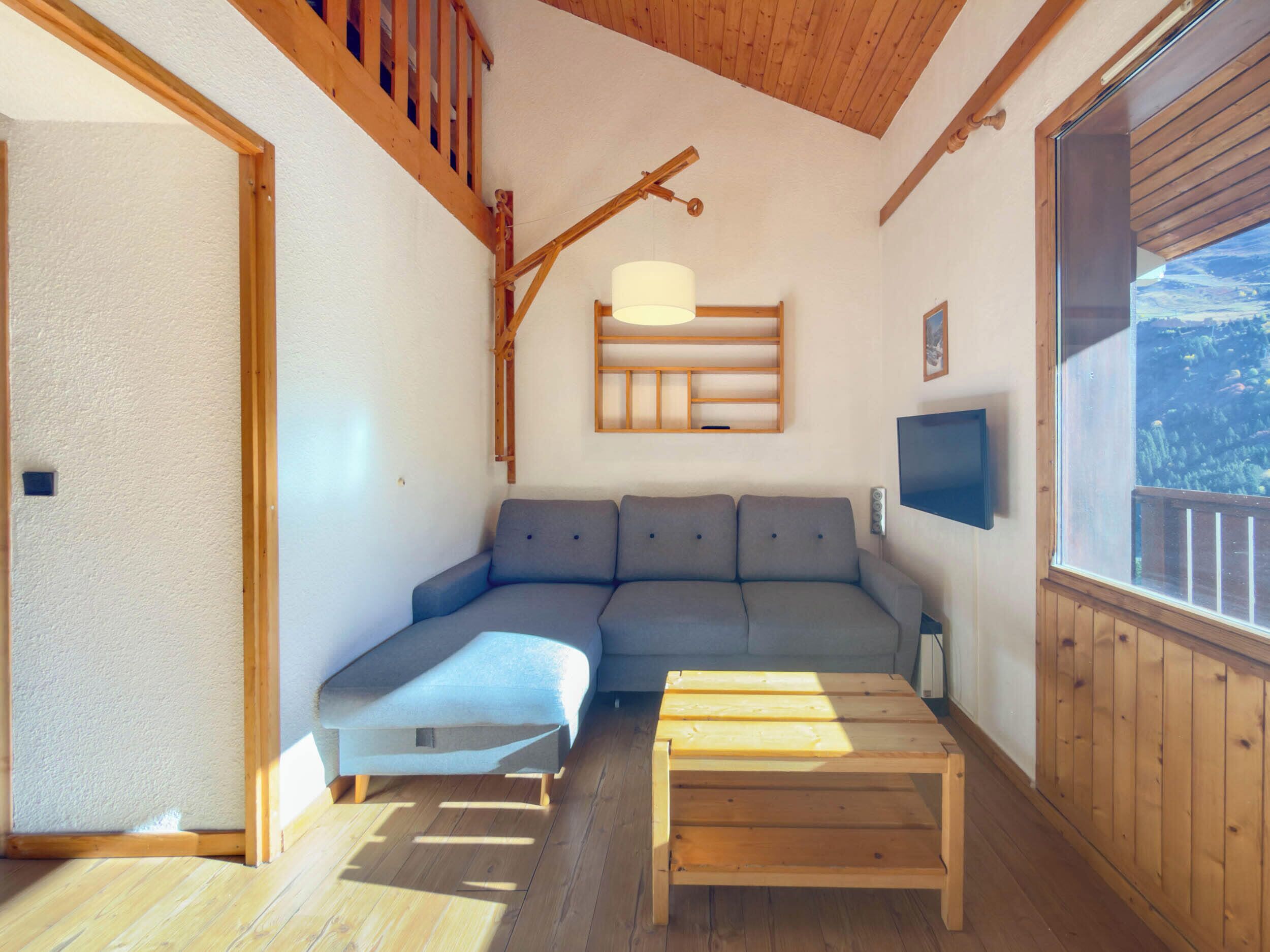 Ein gemütliches Zimmer mit Holzboden, einem grauen Sofa, einem Holztisch und einem an der Wand befestigten Fernseher.