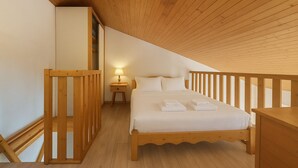 A bedroom with a wooden sloped ceiling, a bed with white linens, a nightstand with a lamp, and a wooden railing.