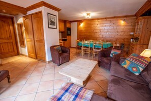Een gezellige woonkamer met houten wanden, een eethoek met een groen tafelkleed, en twee bruine banken met kleurrijke kussens.