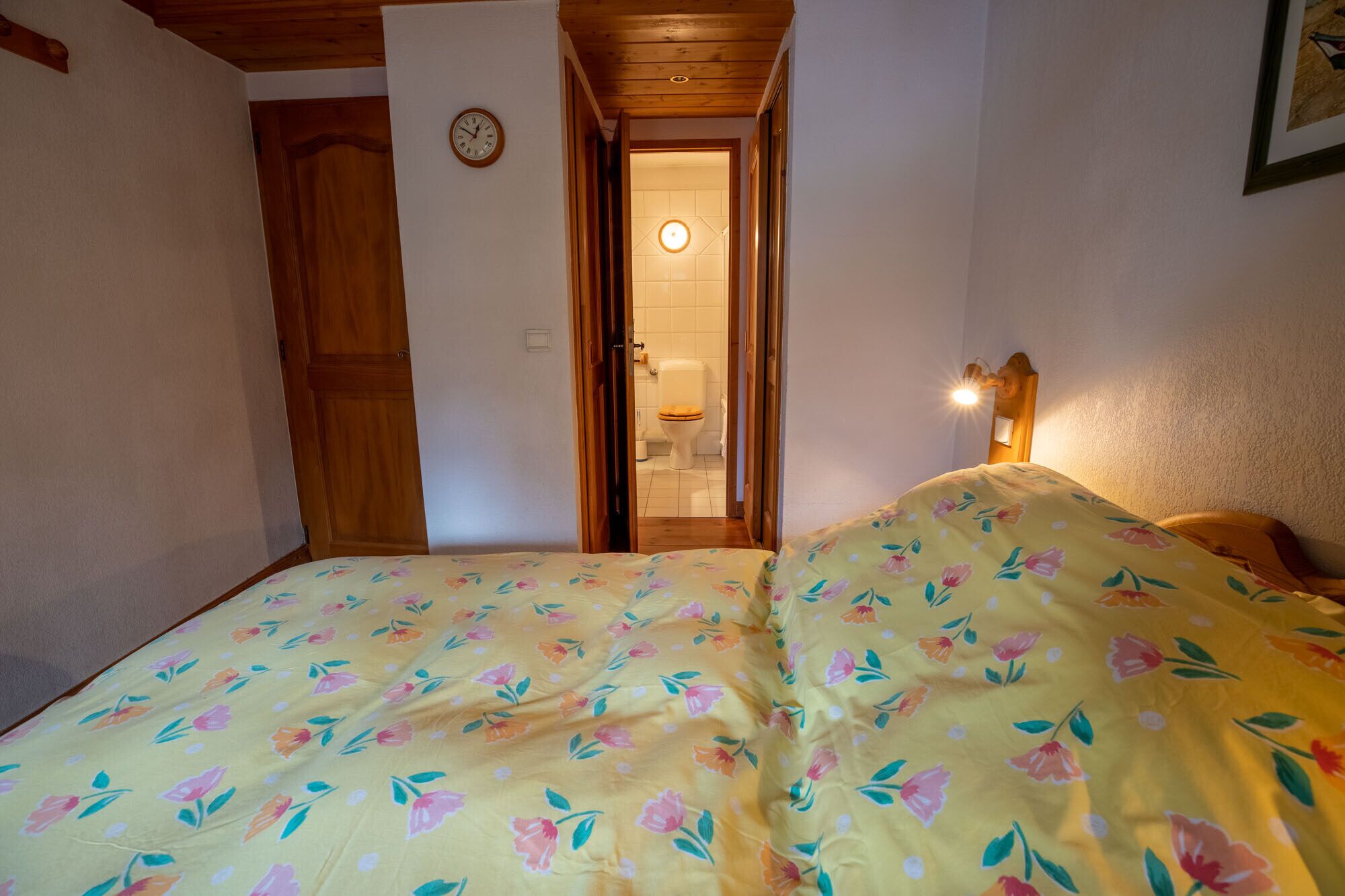 Une chambre à coucher avec un lit, une porte en bois, une horloge et une salle de bain visible à travers une porte ouverte.