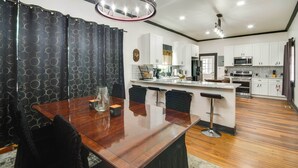 A modern kitchen with white cabinets, a central island, and a dining area with a wooden table and black chairs. - Updated Cottage w/ Fireplaces in North Greenwood! (Greenwood)