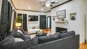 A modern living room with a fireplace, dark grey sofas, a ceiling fan, and a TV mounted on the wall. - Updated Cottage w/ Fireplaces in North Greenwood! (Greenwood)