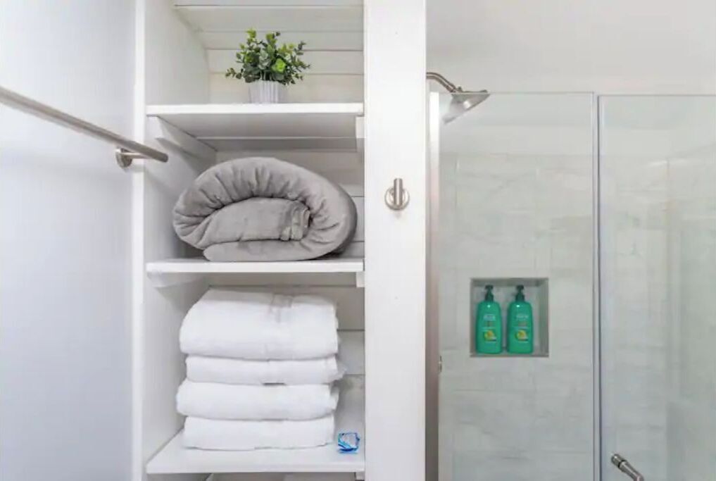 A bathroom with a glass shower enclosure, white shelves with folded towels, and two green bottles on the shelf.