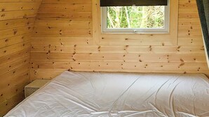 Um pequeno quarto de cabana de madeira, com uma cama, uma janela e vista para o verde lá fora.