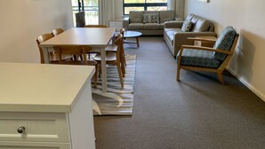 Ein Wohnzimmer mit einer Couch, einem Esstisch mit Stühlen, einem gemusterten Teppich und Blick ins Freie durch große Fenster.