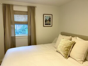 A bedroom with a bed, beige curtains, a window with a view of greenery, and a framed picture on the wall. - Charming Southern Cottage (Savannah)