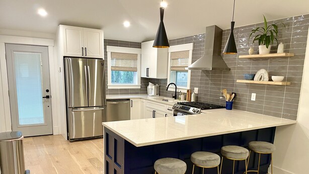 A modern kitchen with stainless steel appliances, a large island with bar stools, and a backsplash of gray tiles. - Charming Southern Cottage (Savannah)