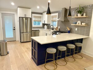 A modern kitchen with stainless steel appliances, a large island with bar stools, and a backsplash of gray tiles. - Charming Southern Cottage (Savannah)