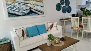 A living room with a white sofa, blue cushions, a coffee table, and a painting of boats on the wall. - Cozy 2BR Apt, 50 meters from the beach with large pool (Progreso)