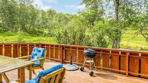 Eine Holzterrasse mit Tisch, Stühlen, einem Grill und Blick auf eine grüne Landschaft.