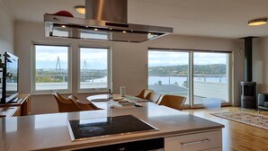 A modern kitchen with a central island, large windows with a view of a bridge and hills, and a wood-burning stove.