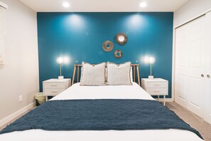 Une chambre à coucher comprenant un lit, deux tables de chevet, une commode, un mur d’accent bleu et des œuvres d’art décoratives.