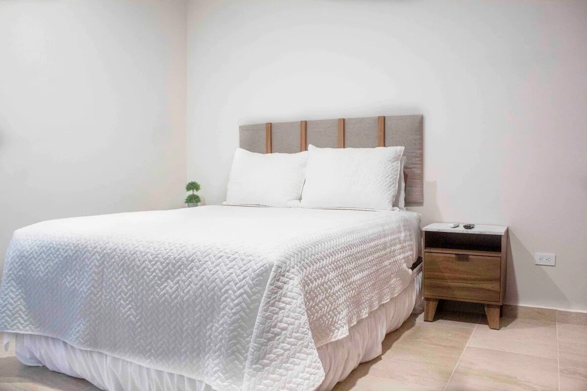 A neatly made bed with a white quilt, a wooden headboard, and a small wooden nightstand beside it.