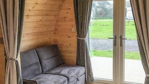 A wooden cabin with a grey sofa and curtains, a sliding glass door, and a view of a grassy area. - Lavender is a cosy pod on the site of the Centry Touring Park in Brixham. (Brixham)